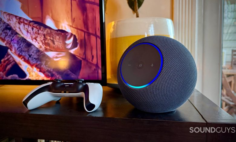 Photo of an Amazon Echo Studio on a black wooden surface next to a PS5 controller with a TV fireplace on in the background.