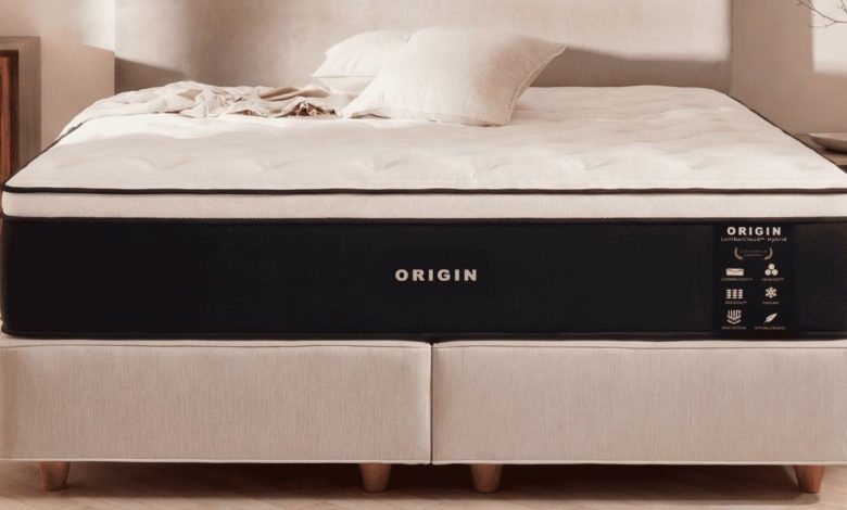 A professionally staged studio shot showcases the Origin LumbarCloud Hybrid mattress on a modern divan base in a bright, minimalist bedroom.