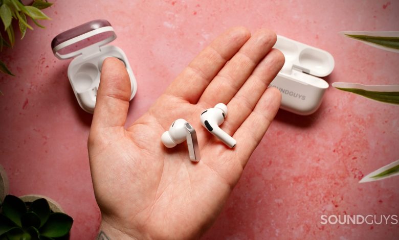 The Samsung Galaxy Buds 4 Pro and Apple AirPods Pro (3rd generation) earbuds resting in a hand over a pink textured surface with their charging cases in the background.