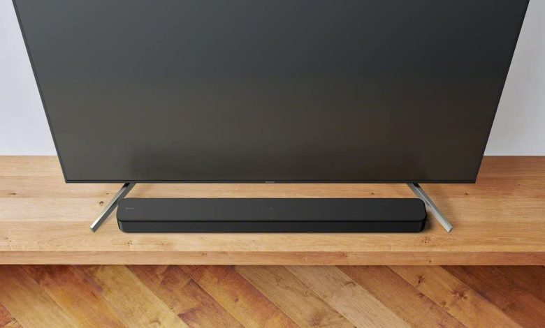 The Sony HT-SF150 soundbar positioned on a wooden cabinet, between the feet of a TV