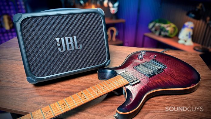 The JBL BandBox Trio on a wooden table in the studio with a PRS guitar next to it.