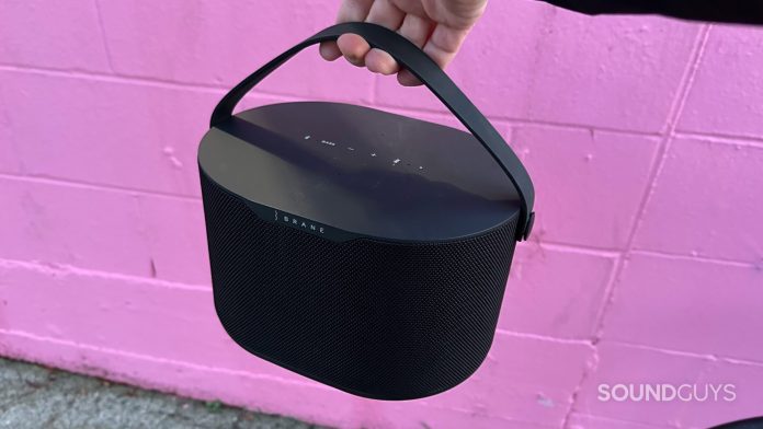 A hand holding the Brane X speaker by the handle in front of a pink building.