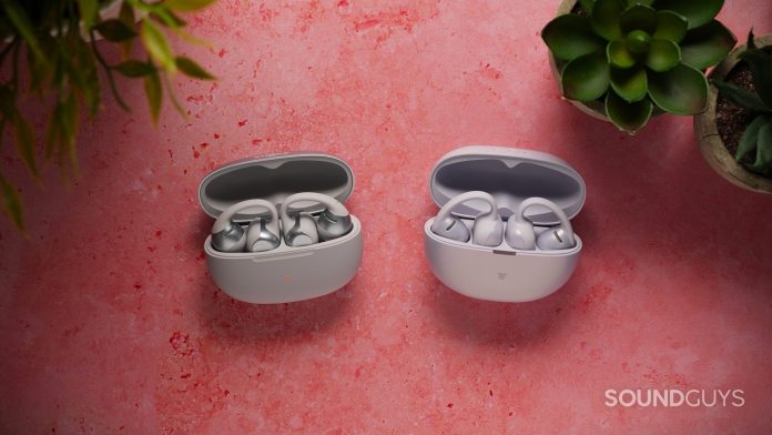 The Shokz OpenDots ONE and OpenDots Air sit inside their open charging cases on a pink tabletop. The earbuds are arranged for a side-by-side comparison, with small potted succulents positioned around the scene.
